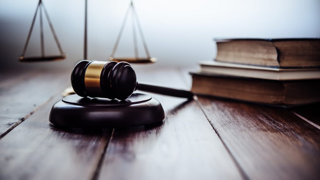 A portrait of a hammer and gavel, with old books off to the right and a scale of justice in the back left corner.