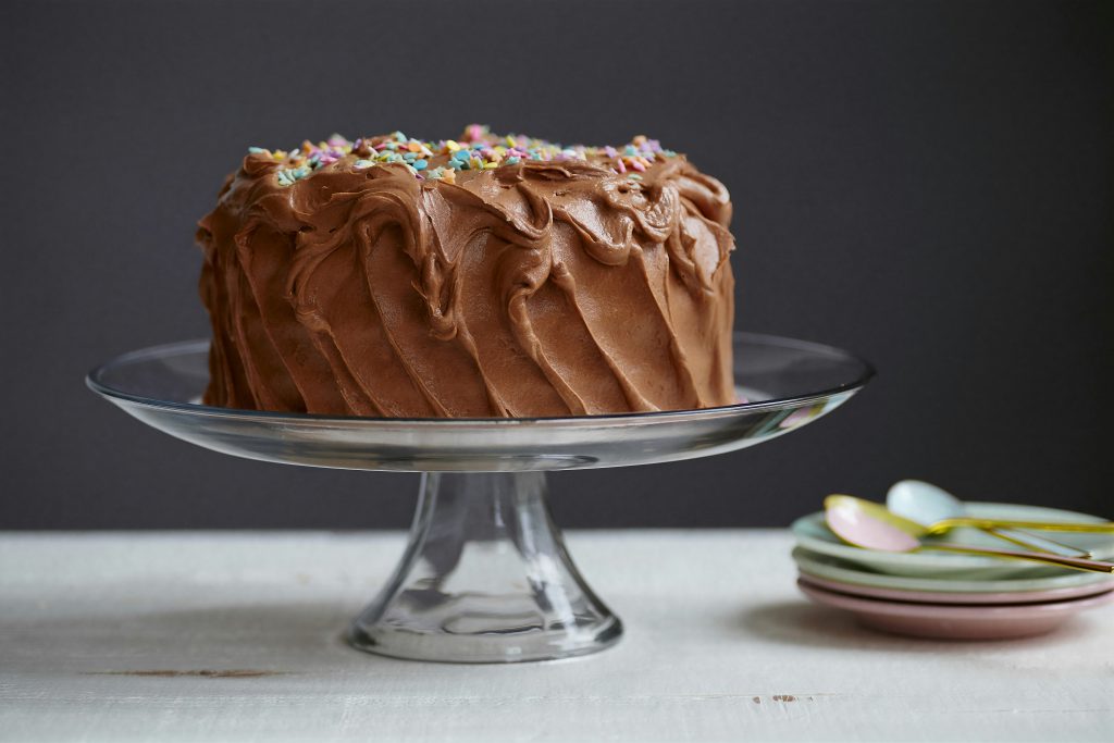 Chocolate cake with pastel jimmies on a glass cake platter, with teaspoons and plates in the background