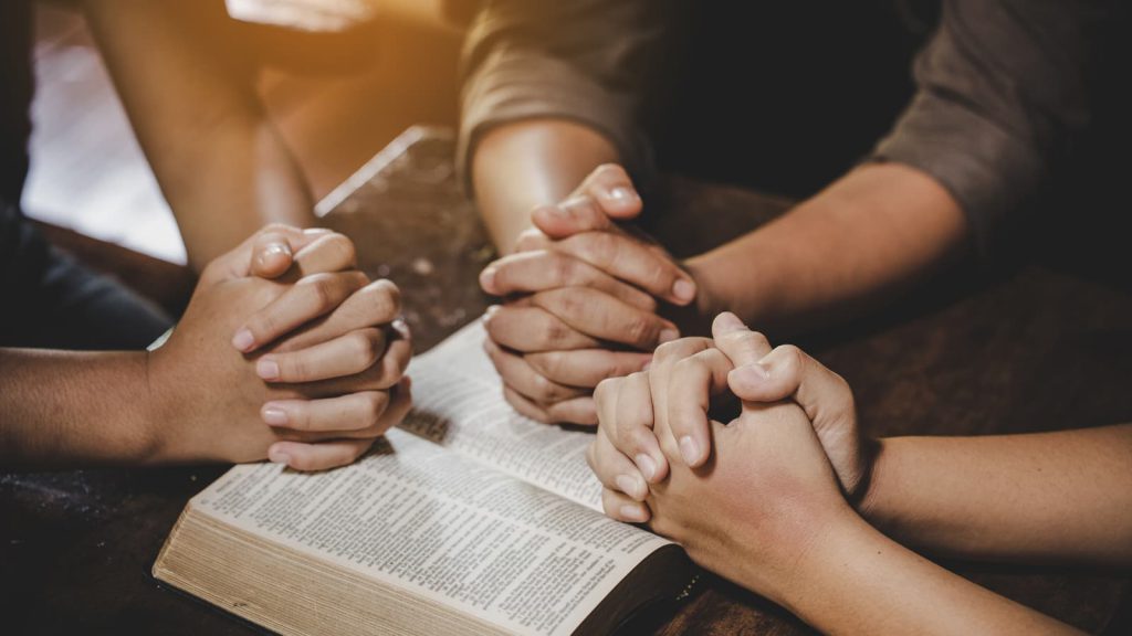 Three sets of hands folded in prayer over an open Bible
